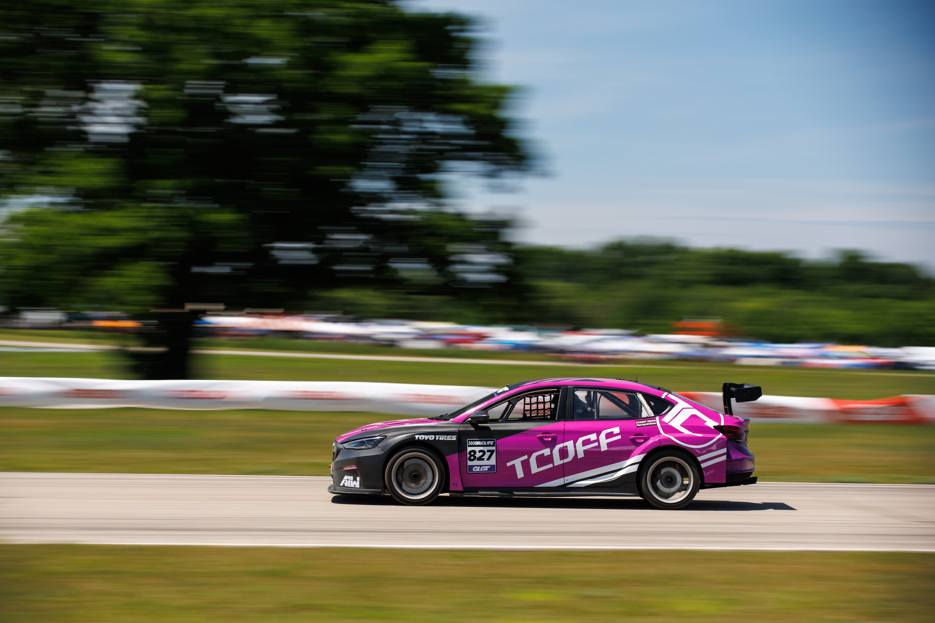 A purple and black MG6 TCR race car with 'TCOFF', captured with a motion-blurred background of trees and track barriers.